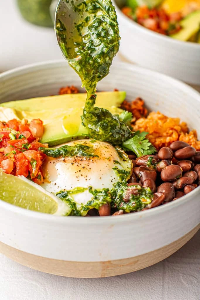 A vibrant green sauce being drizzled over a fried egg and avocado in the bowl